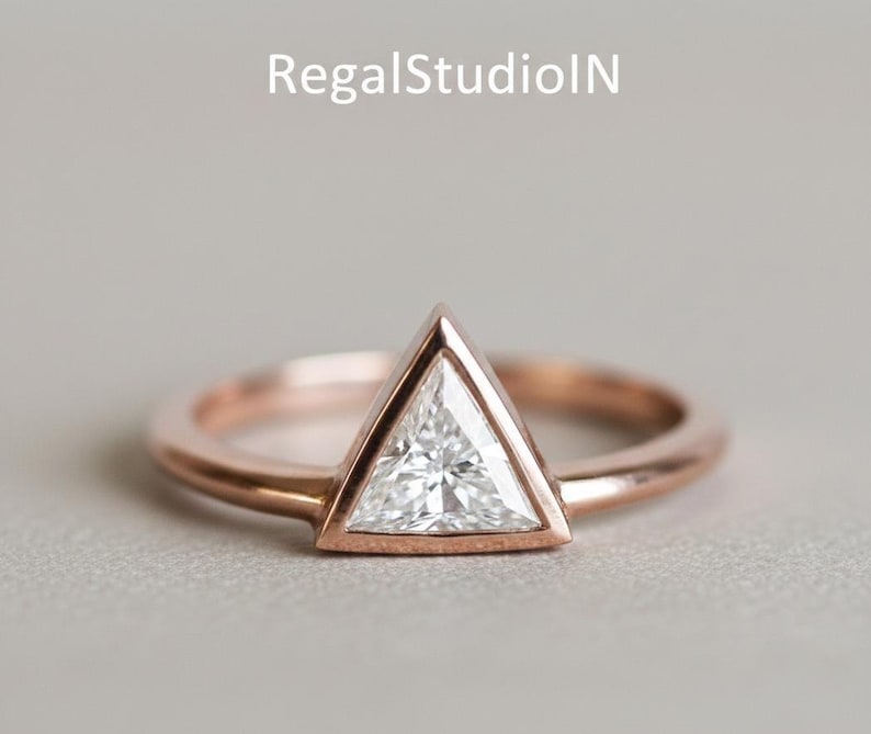 Peut inclure: Une bague en or rose avec une pierre pr&eacute;cieuse blanche triangulaire. La pierre est sertie dans un sertissage &agrave; lunette, et le bracelet est lisse et poli. La bague est pr&eacute;sent&eacute;e sur une surface grise neutre.