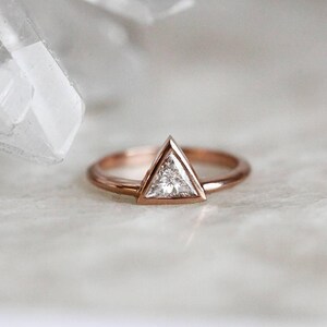 Peut inclure: Bague en or rose avec un diamant triangulaire. Le diamant est serti dans un sertissage &agrave; lunette, et le bracelet est poli. La bague est pr&eacute;sent&eacute;e sur une surface blanche. Le design de la bague est simple et &eacute;l&eacute;gant.