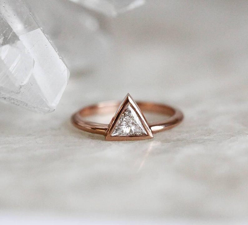 Peut inclure: Bague en or rose avec un diamant triangulaire. Le diamant est serti dans un sertissage &agrave; lunette, et le bracelet est poli. La bague est pr&eacute;sent&eacute;e sur une surface blanche. Le design de la bague est simple et &eacute;l&eacute;gant.