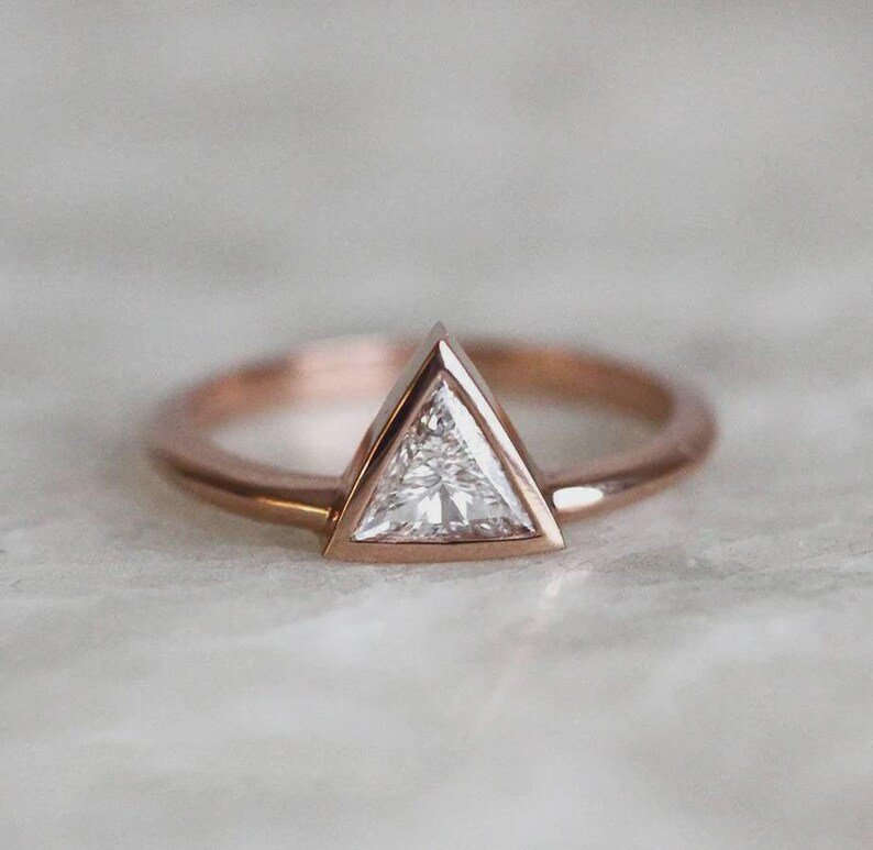 Peut inclure: Bague en or rose avec un diamant taill&eacute; en triangle. Le diamant est serti dans un sertissage &agrave; lunette, et le bracelet est en or rose fin et poli. La bague est pr&eacute;sent&eacute;e sur une surface de couleur claire.