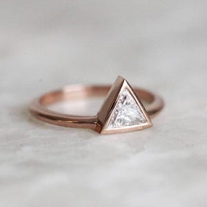 Peut inclure: Une bague en or rose orn&eacute;e d'une pierre pr&eacute;cieuse triangulaire et transparente. La pierre est sertie dans un sertissage &agrave; lunette, et le corps de la bague est de conception simple et polie. La bague est pr&eacute;sent&eacute;e sur une surface claire.