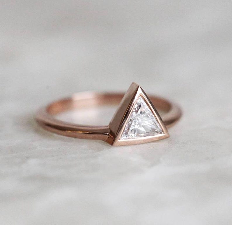 Peut inclure: Une bague en or rose orn&eacute;e d'une pierre pr&eacute;cieuse triangulaire et transparente. La pierre est sertie dans un sertissage &agrave; lunette, et le corps de la bague est de conception simple et polie. La bague est pr&eacute;sent&eacute;e sur une surface claire.