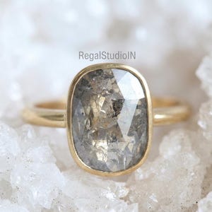 May include: A gold ring with a large, rectangular, salt and pepper diamond. The diamond is set in a gold bezel setting. The ring is displayed on a white, crystalline surface. The text "RegalstudioIN" is visible.