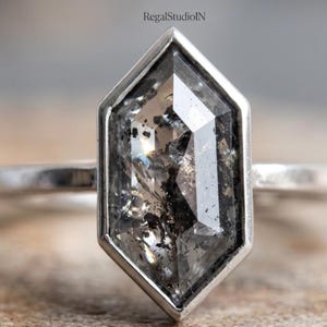 May include: A silver ring featuring a large, faceted, hexagonal-shaped gemstone with gray and black inclusions. The gemstone is set in a silver bezel. The ring is displayed on a wooden surface. The text "RegalStudioIN" is in the upper right corner.