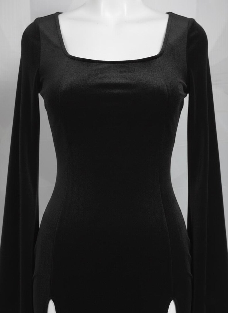 Op de afbeelding: Een zwarte jurk met lange mouwen en een vierkante halslijn. De jurk heeft een getailleerd lijfje en een split aan de zoom. De stof lijkt een zacht, fluweelachtig materiaal te zijn. De jurk wordt op een mannequin getoond.