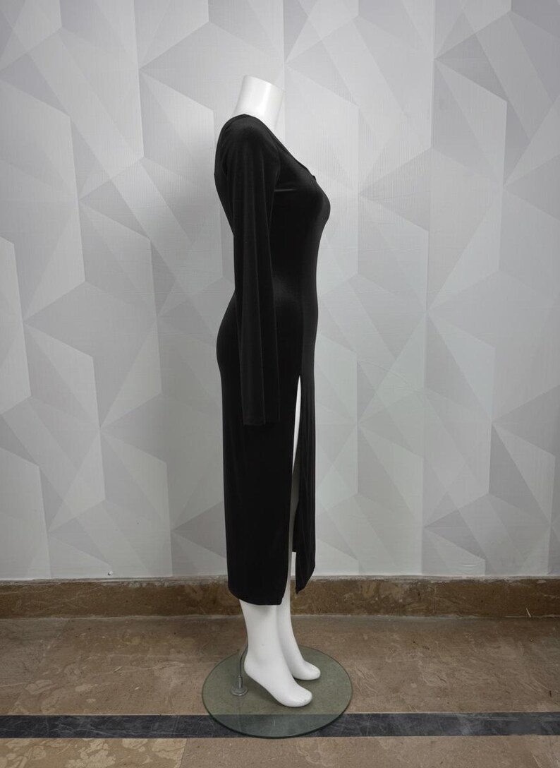 Op de afbeelding: Een lange, zwarte jurk met lange mouwen en een hoge split aan de linkerkant. De jurk wordt getoond op een mannequin met witte voeten en een witte kop. De jurk heeft een V-hals.