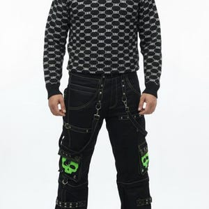 Puede incluir: Una persona con un jersey negro de cuello alto con un patrón blanco y pantalones negros. Los pantalones tienen parches de calaveras verdes y detalles de cadena plateada. El atuendo tiene una estética gótica o punk.