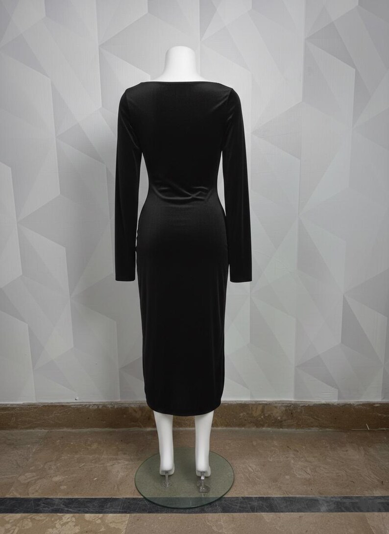 Op de afbeelding: Een lange zwarte jurk met lange mouwen en een zoom tot op de knie. De jurk heeft een ronde hals en een getailleerd silhouet. De jurk wordt gepresenteerd op een mannequin tegen een witte geometrisch patroon muur.
