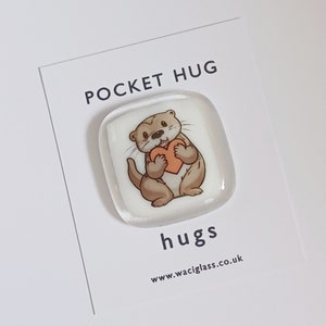May include: A white glass pocket hug with a cartoon otter holding a heart. The glass is square and has a rounded edge. The text "POCKET HUG" is printed on the card behind the glass. The text "hugs" is printed below the glass. The text "www.waciglass.co.uk" is printed below the text "hugs".