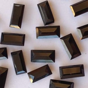 May include: A scattering of rectangular, black gemstones on a white background. The gemstones have a polished, reflective surface and a faceted cut. The gems are of varying sizes and orientations, creating a visually interesting arrangement.