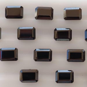 May include: A selection of rectangular, black gemstones with a polished finish. The gems are arranged in rows, each with a symmetrical, emerald-cut shape. The dark colour of the stones contrasts with the light background.