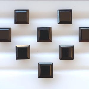 May include: Nine square-cut, black gemstones on a white surface. Each gem has a polished, reflective surface and a square shape. The gems are evenly spaced, creating a pattern. The gemstones are likely for jewelry making, each approximately 1 cm in size.