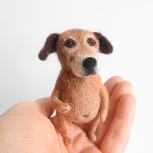 May include: A small, brown and white needle felted dog with brown ears. The dog is sitting on a person's hand.