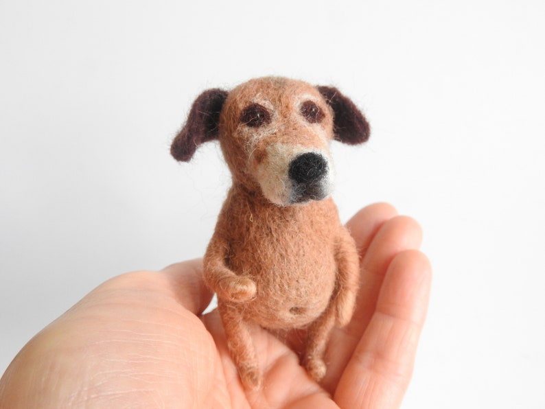May include: A small, brown and white needle felted dog with brown ears. The dog is sitting on a person's hand.
