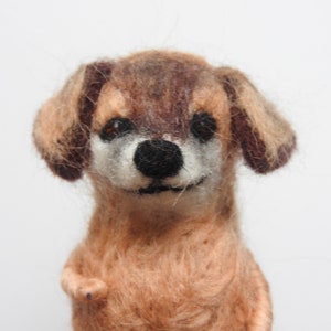May include: A brown and tan needle felted dog with brown eyes and a black nose. The dog is smiling and has a fluffy texture.