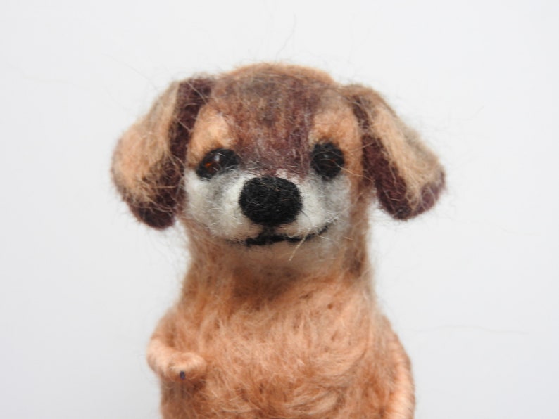 May include: A brown and tan needle felted dog with brown eyes and a black nose. The dog is smiling and has a fluffy texture.