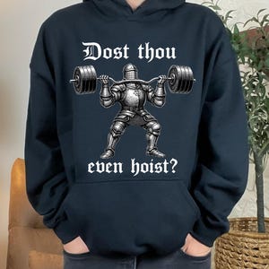 K&ouml;nnte beinhalten: Schwarzer Kapuzenpullover mit einem Gewichtheber-Ritter-Motiv. Der Ritter, in R&uuml;stung, hebt eine Langhantel. Der Text "Dost thou even hoist?" ist in wei&szlig;er gotischer Schrift &uuml;ber und unter dem Ritter gedruckt.