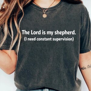May include: Dark grey t-shirt with the text "The Lord is my shepherd. (I need constant supervision)". The shirt is a short-sleeved crew neck style.