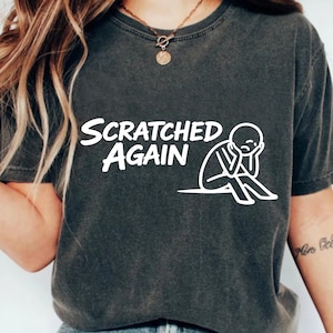 Pode incluir: T-shirt cinzenta escura com as palavras brancas "SCRATCHED AGAIN" e uma ilustração branca de uma pessoa sentada com a cabeça nas mãos. A t-shirt é de estilo gola redonda.
