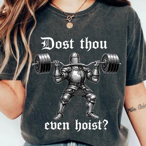 K&ouml;nnte beinhalten: Dunkelgraues T-Shirt mit einer Schwarz-Wei&szlig;-Illustration eines Ritters, der eine Langhantel hebt. Der Text "Dost thou even hoist?" ist in wei&szlig;er, gotischer Schrift gedruckt. Ein l&auml;ssiges Kleidungsst&uuml;ck.