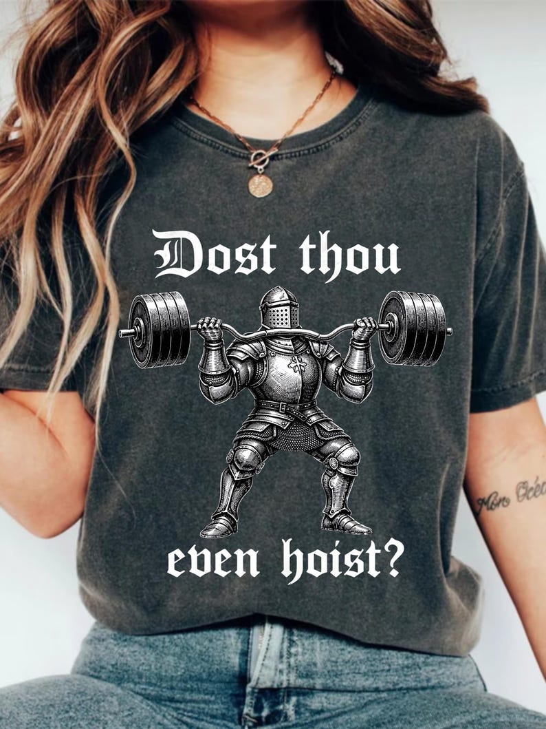 K&ouml;nnte beinhalten: Dunkelgraues T-Shirt mit einer Schwarz-Wei&szlig;-Illustration eines Ritters, der eine Langhantel hebt. Der Text "Dost thou even hoist?" ist in wei&szlig;er, gotischer Schrift gedruckt. Ein l&auml;ssiges Kleidungsst&uuml;ck.
