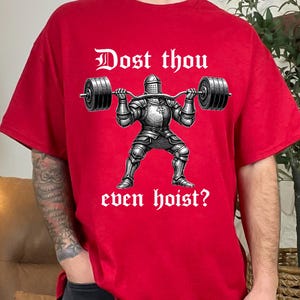 K&ouml;nnte beinhalten: Rotes T-Shirt mit einer Grafik eines Ritters, der eine Langhantel hebt. Der Text "Dost thou even hoist?" ist oberhalb und unterhalb der Ritterillustration in gotischer Schrift gedruckt. Das Shirt ist aus weichem Material.