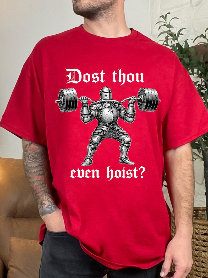 K&ouml;nnte beinhalten: Rotes T-Shirt mit einer Grafik eines Ritters, der eine Langhantel hebt. Der Text "Dost thou even hoist?" ist oberhalb und unterhalb der Ritterillustration in gotischer Schrift gedruckt. Das Shirt ist aus weichem Material.