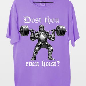 K&ouml;nnte beinhalten: Ein lilafarbenes T-Shirt mit einer Grafik eines Ritters, der eine Langhantel hebt. Der Text "Dost thou even hoist?" ist in gotischer Schrift &uuml;ber und unter dem Ritter aufgedruckt. Das T-Shirt h&auml;ngt an einem Holzkleiderb&uuml;gel.