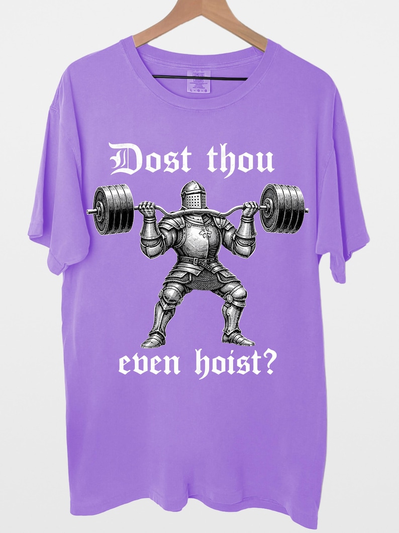 K&ouml;nnte beinhalten: Ein lilafarbenes T-Shirt mit einer Grafik eines Ritters, der eine Langhantel hebt. Der Text "Dost thou even hoist?" ist in gotischer Schrift &uuml;ber und unter dem Ritter aufgedruckt. Das T-Shirt h&auml;ngt an einem Holzkleiderb&uuml;gel.
