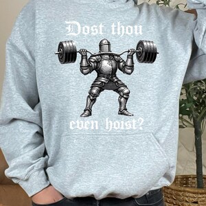 K&ouml;nnte beinhalten: Hellgrauer Kapuzenpullover mit dem Text "Dost thou even hoist?" &uuml;ber einer Schwarz-Wei&szlig;-Illustration eines Ritters, der Gewichte hebt. Der Ritter tr&auml;gt eine volle R&uuml;stung und h&auml;lt eine Langhantel.