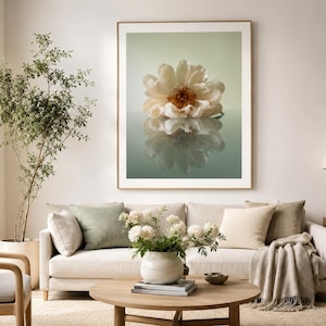 Puede incluir: Una impresión enmarcada de una flor grande y clara con un centro marrón, reflejada en una superficie brillante. La obra de arte se exhibe sobre un sofá beige con cojines verdes y crema. Un jarrón de flores blancas se encuentra sobre una mesa de centro de madera.