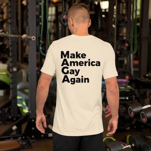 Könnte beinhalten: Cremefarbenes T-Shirt mit dem Text "Make America Gay Again" in fetten schwarzen Buchstaben. Das kurzärmelige Shirt scheint aus einem weichen Material zu sein. Die Person befindet sich in einem Fitnessstudio.
