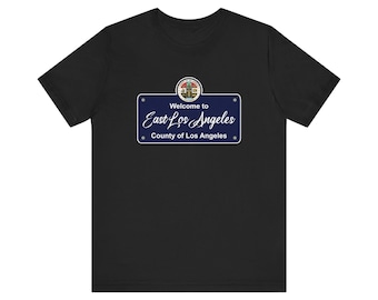 May include: Black t-shirt featuring a blue sign design with the text "Welcome to East Los Angeles County of Los Angeles." The sign has a white border and a circular emblem at the top.