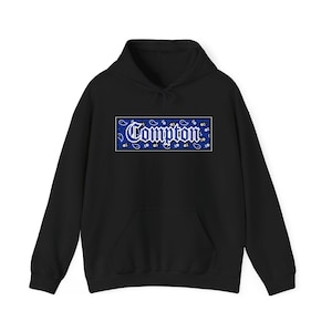 Puede incluir: Sudadera con capucha negra con bolsillo delantero y cordón. La sudadera presenta un diseño rectangular azul con la palabra "Compton" en escritura blanca, rodeada de un estampado de cachemira.