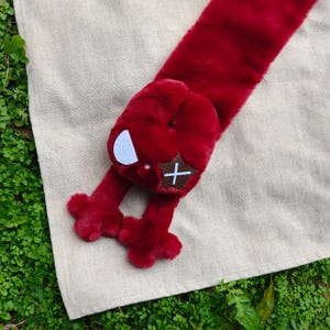 Op de afbeelding: Een rode pluche sjaal met een speels ontwerp. De sjaal heeft een grote, afgeronde kop met een witte halve maan en een bruine patch met een wit kruis. Twee rode, geklauwde voeten steken uit de kop. De sjaal ligt op een beige ondergrond.