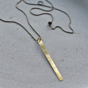 Op de afbeelding: Een lange, dunne, gouden staafhanger hangt aan een delicate ketting. De hanger heeft een gehamerde textuur en een licht onregelmatige vorm.