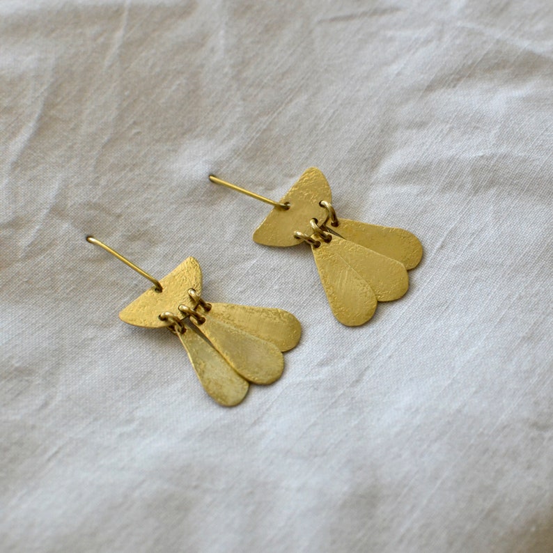 May include: A pair of gold earrings with a geometric design. Each earring features a small, curved top piece with three teardrop-shaped pendants hanging below.