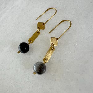 K&ouml;nnte beinhalten: Ein Paar goldene Ohrringe mit einem strukturierten quadratischen Anh&auml;nger und einer grauen Steinperle an jedem Ohrring.