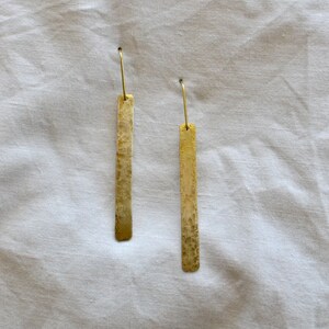 May include: A pair of gold-toned drop earrings featuring long, rectangular metal bars with a textured, mottled surface finish. Each earring is suspended from a thin, curved wire hook, creating a simple, elongated silhouette with a metallic sheen.