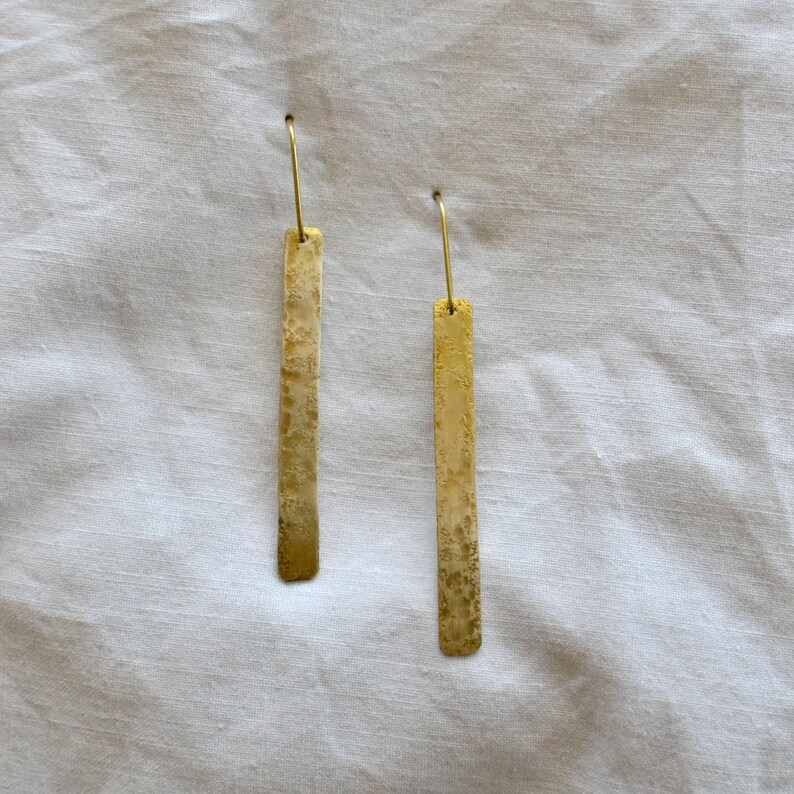 May include: A pair of gold-toned drop earrings featuring long, rectangular metal bars with a textured, mottled surface finish. Each earring is suspended from a thin, curved wire hook, creating a simple, elongated silhouette with a metallic sheen.