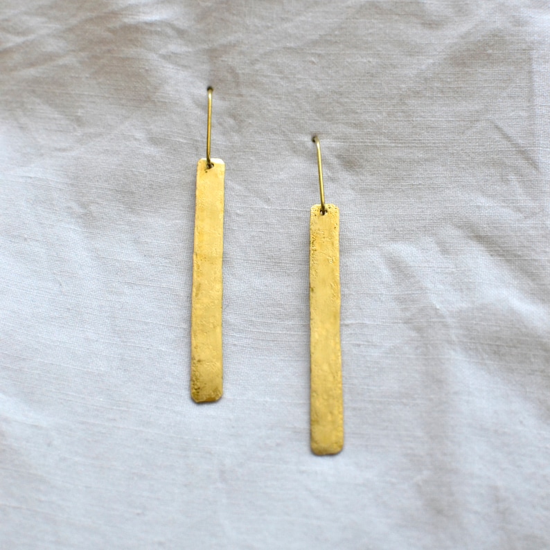 May include: A pair of gold-toned drop earrings featuring long, rectangular metal bars with a textured, hammered finish. Each earring is suspended from a thin, curved metal hook, creating a simple, elongated silhouette with a metallic luster.
