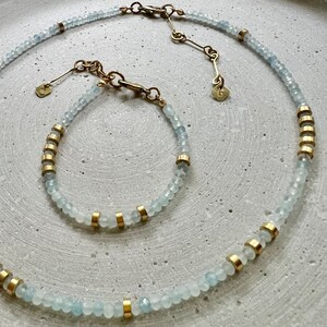 May include: A necklace and bracelet set featuring faceted pale blue beads interspersed with small gold-toned cylindrical spacers. Both pieces include a gold-toned metal clasp, an adjustable chain, and a small square tag with the letter G engraved.