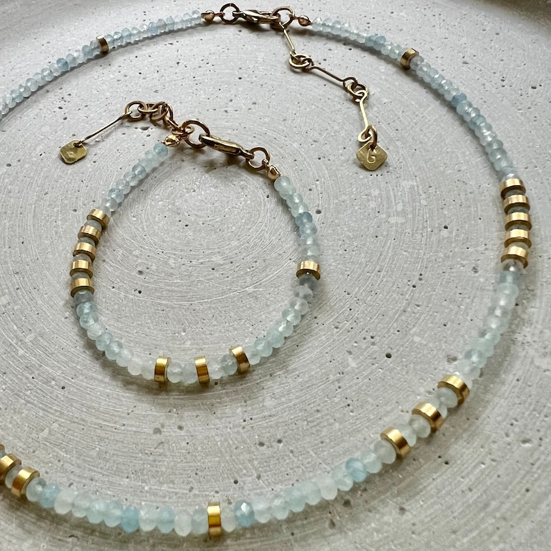 May include: A necklace and bracelet set featuring faceted pale blue beads interspersed with small gold-toned cylindrical spacers. Both pieces include a gold-toned metal clasp, an adjustable chain, and a small square tag with the letter G engraved.