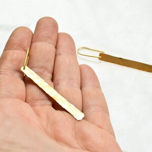 May include: A pair of gold-toned earrings featuring long, thin, rectangular metal bars with a hammered, textured finish. Each bar is attached to a simple, curved gold-toned ear wire hook, creating a dangling, linear design that hangs vertically.