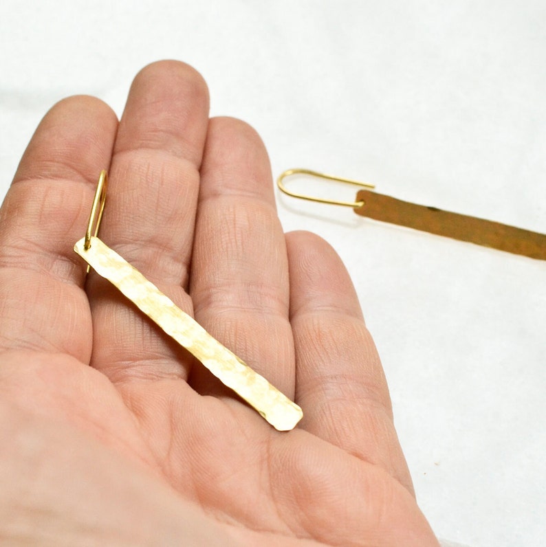 May include: A pair of gold-toned earrings featuring long, thin, rectangular metal bars with a hammered, textured finish. Each bar is attached to a simple, curved gold-toned ear wire hook, creating a dangling, linear design that hangs vertically.