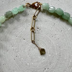 May include: A close-up of a necklace with light green, oval beads. The necklace has a gold-coloured clasp and chain. The beads are strung together, creating a delicate accessory. The necklace is on a grey surface.