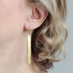 May include: A long, thin, gold-toned metal earring with a hammered, textured surface. The earring features a slender rectangular bar shape suspended from a simple, curved wire hook that passes through the earlobe, creating a vertical drop design.