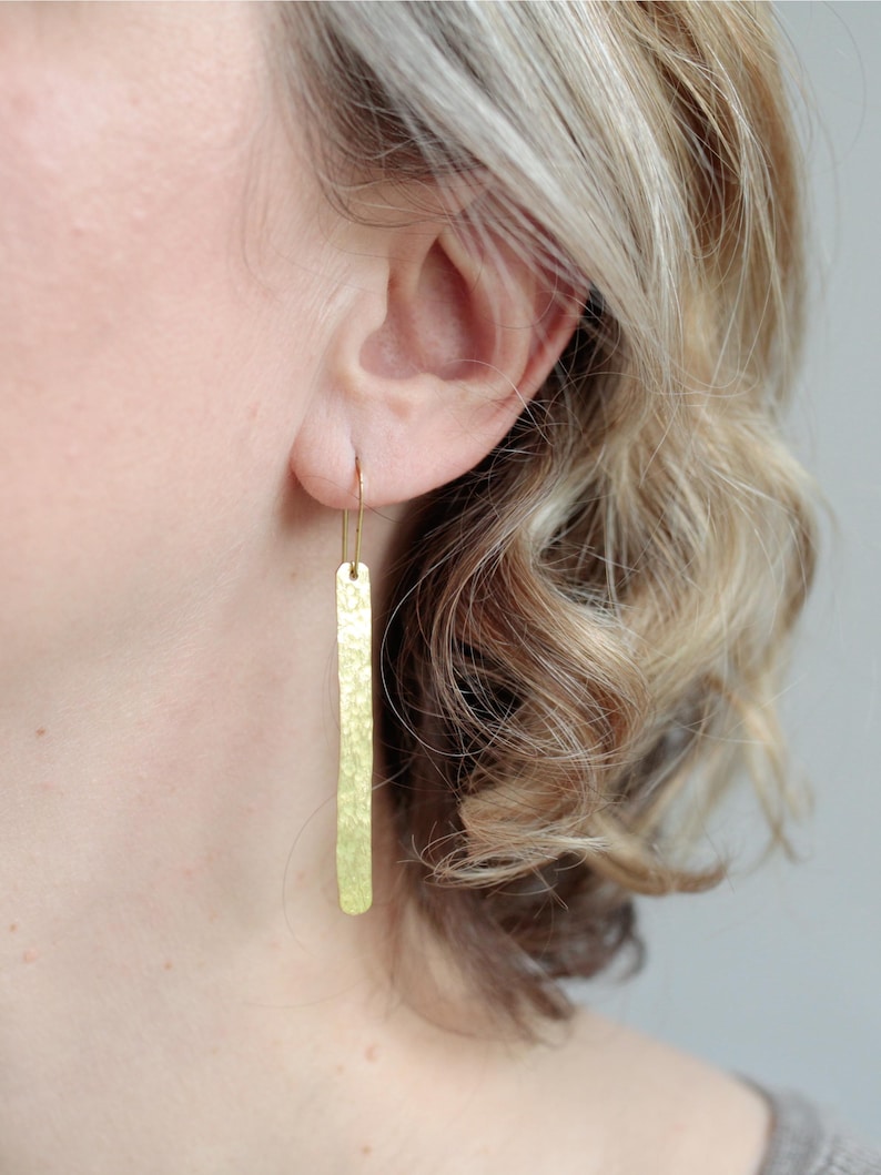 May include: A long, thin, gold-toned metal earring with a hammered, textured surface. The earring features a slender rectangular bar shape suspended from a simple, curved wire hook that passes through the earlobe, creating a vertical drop design.