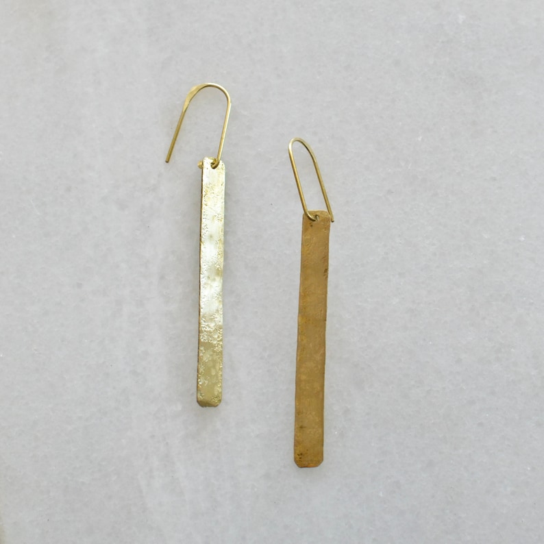 May include: A pair of gold-toned metal earrings featuring long, slender rectangular bars with a textured, hammered finish. Each earring is suspended from a simple, curved wire hook that attaches to the top of the vertical bar, creating a drop design.