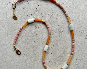 May include: A beaded necklace featuring a mix of pink, orange, white, and green beads. The necklace has a gold-colored clasp and a small tag. The beads are arranged in a linear pattern, creating a colorful and stylish accessory.